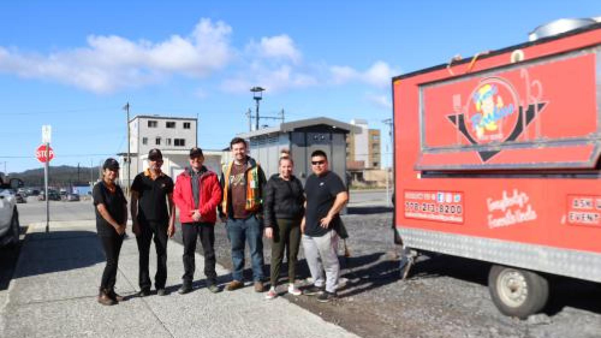 A red food truck is parked next to 5 people lined up for a photo in a parking lot. 