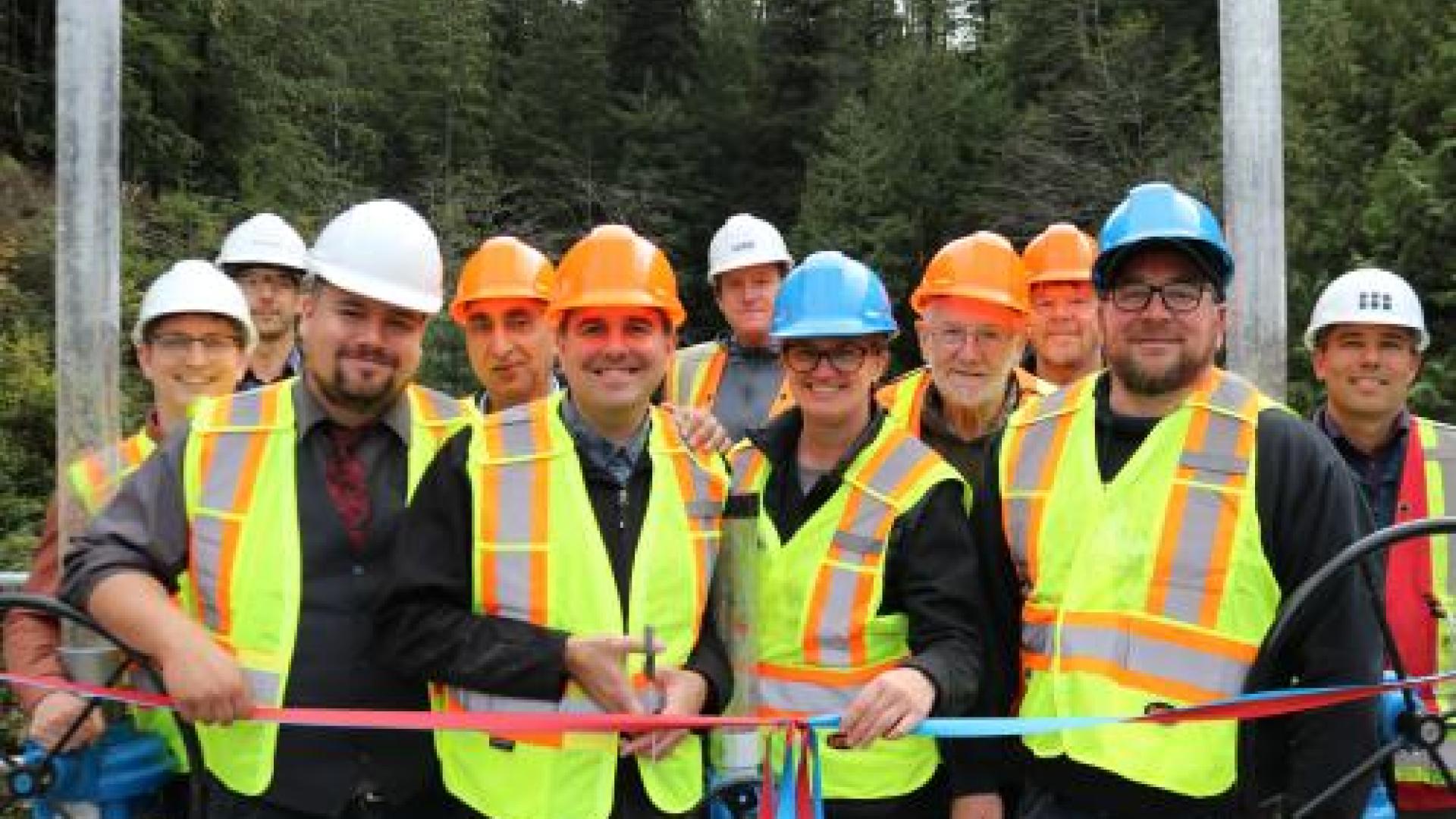 Several adults in high visibility vests and hard hats prepare to cut a ribbon. 