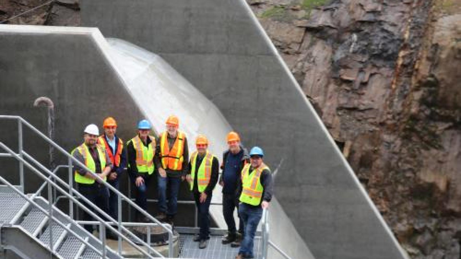 Seven adults stand in a line at a distance, in front of the newly built dam.