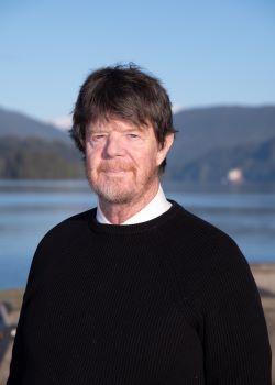 Headshot of Councillor Nick Adey. There is a man with brown hair and facial hair wearing a black sweater with a neutral facial expression. The ocean and mountains are in the background.