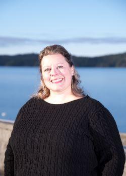 Headshot of Councillor Teri Forster. There is a woman smiling wearing a black dress. The ocean is in the background.