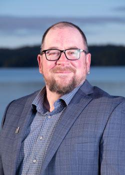Headshot of Councillor Wade Niesh. There is a man with short hair, facial hair, and glasses wearing a navy sports jacket and grey shirt smiling. The ocean is in the background.