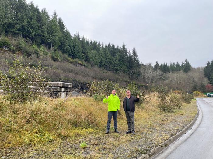 Two people standing on plot of land waving and smiling. The person on the left is wearing a lime green jacket while the person on the right is wearing a black jacket. There is hilly terrain in the background with tall trees on them.